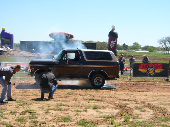 Brown 1978 Ford Bronco doing a burnout, kicking up smoke. Two people are near the front tire, one crouching. A large Dr. Pepper bottle and a purple jug are in the background