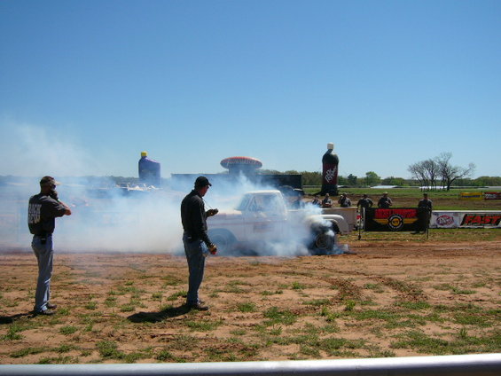 Pickup truck doing a burnout, kicking up a lot of smoke. Two men stand nearby watching