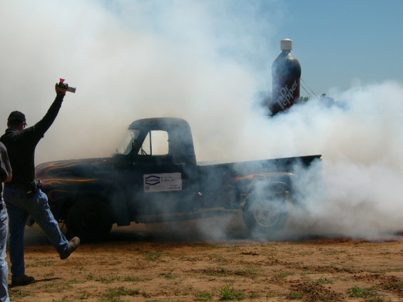 Black pickup truck with flames on the side doing a burnout, emitting smoke. A person is visible on the left