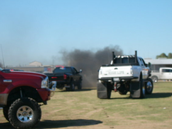 White pickup truck with large tires and exhaust stacks emitting black smoke. Text on tailgate reads 'SMOKE LIFE'