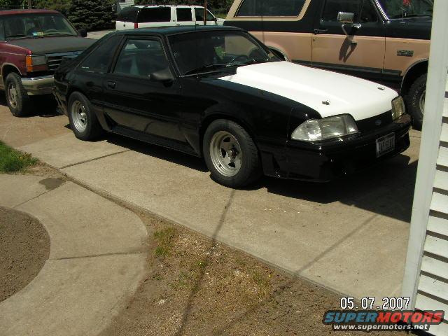 Black 1988 Ford Mustang with white hood, parked on driveway next to a red pickup truck and a brown SUV