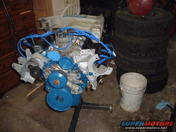 Ford V8 engine, blue spark plug wires, on stand. Stack of tires and bucket nearby