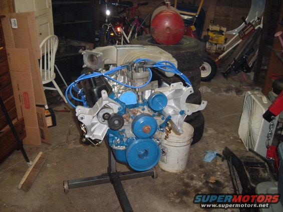 Ford V8 engine on a stand, painted blue and silver, in a garage with tools and tires in background