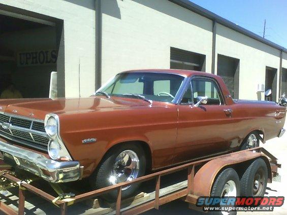 Copper Ford Ranchero on a trailer, front 3/4 view. Chrome wheels and trim. Building in background
