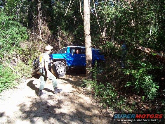 Blue SUV navigating a wooded trail with a person walking nearby