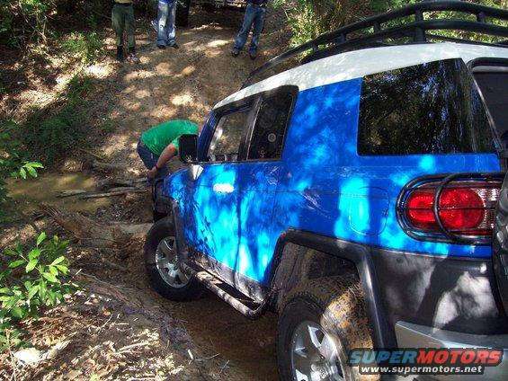 Blue and white SUV stuck in mud on a dirt trail, with people around it