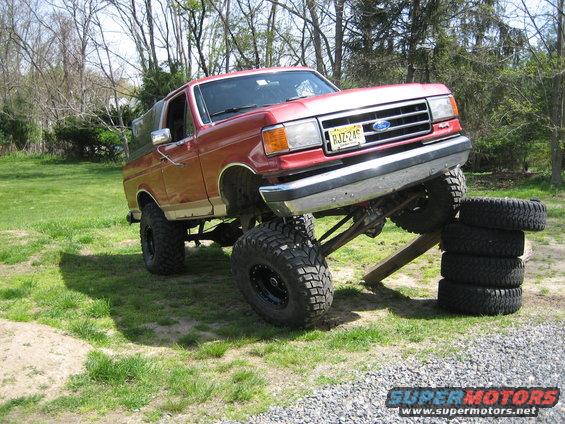 Red lifted Ford Bronco on wooden blocks with tires stacked nearby. License plate RJZ-245
