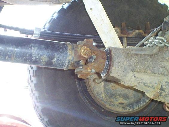 Close-up of a rusty driveshaft connected to a rear axle with a leaf spring and tire visible