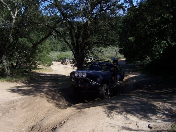 Off-road vehicles navigating a dirt trail under trees. One vehicle is in the foreground, modified for off-roading
