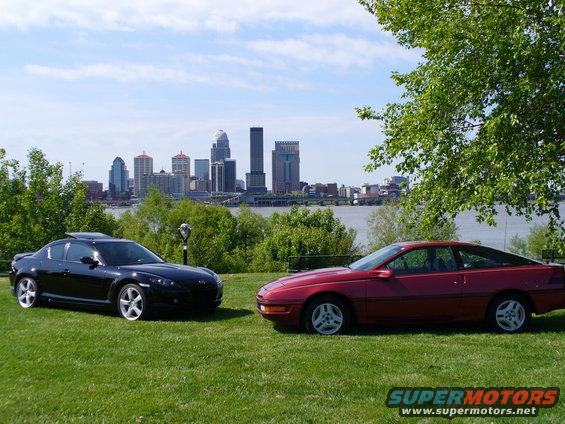 Red 1991 Ford Probe side view with black coupe and city skyline