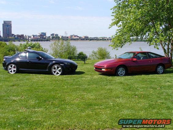 1991 Ford Probe and a black Mazda RX-8 parked on grass by a river with a city skyline in the background