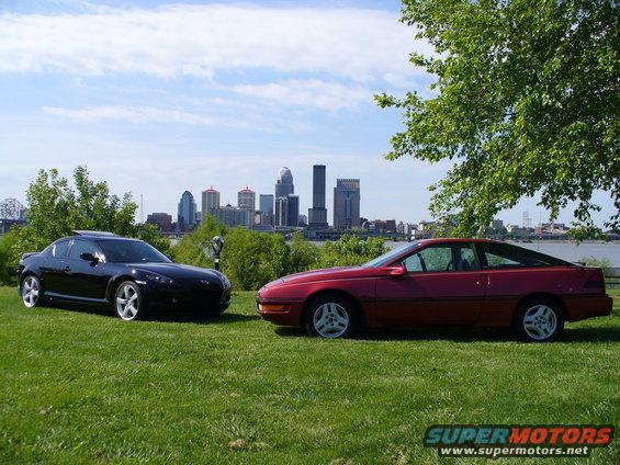 Red sports car and black sports car parked on grass with city skyline in background