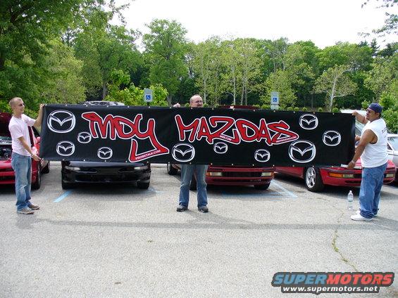 People hold a banner reading 'MDU MAZDAS' in front of several Mazda cars