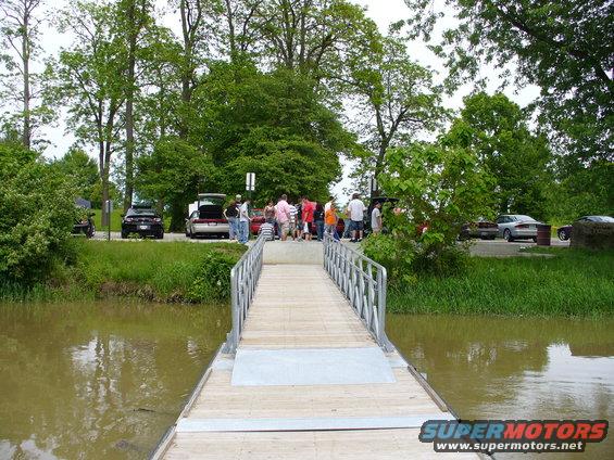 People gathered around cars parked near a river with a wooden walkway in the foreground