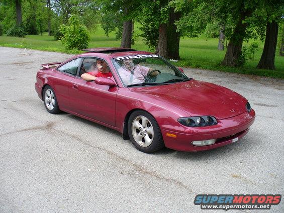 Red Mazda MX-6 with two people inside, parked on asphalt with trees in the background