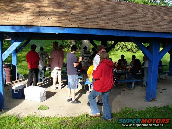 Group of people gathered under a blue picnic shelter, some standing, some sitting at tables