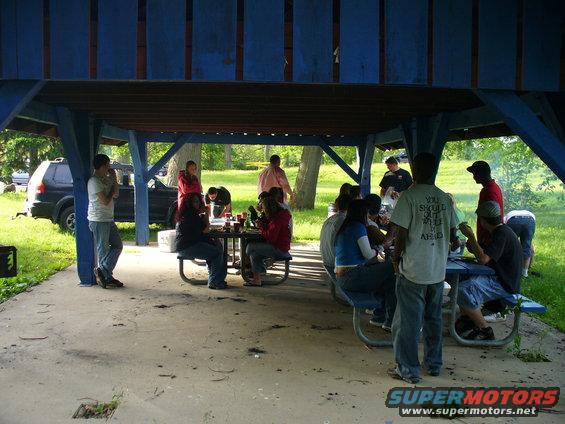 Black SUV parked under a blue pavilion with people gathered at picnic tables