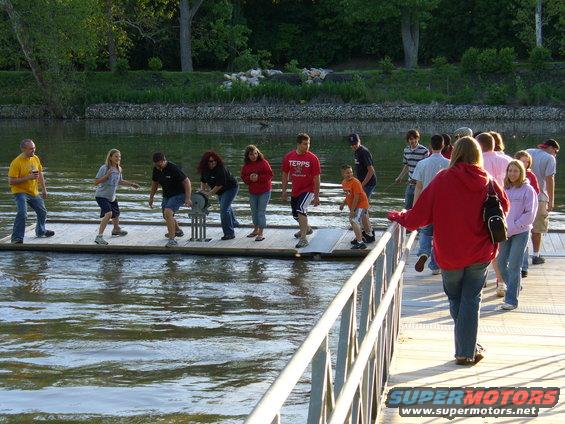 Group of people on a floating dock over water, with trees and a riverbank in the background. A watermark reads SUPERMOTORS