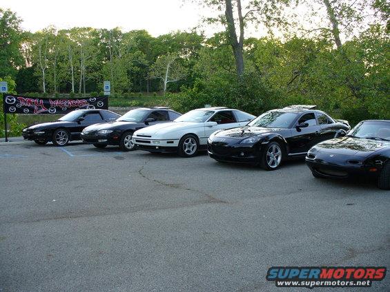 Several Mazda cars parked in a lot, including a white FC RX-7, a black RX-8, and a black Miata