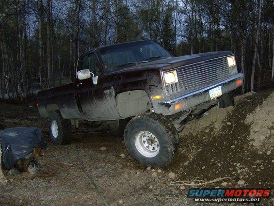 Dark brown pickup truck with a chrome grille, stuck in dirt, with a spare engine covered in plastic to the left