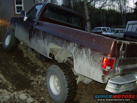 Mud-covered 1982 Chevrolet C/K Pickup truck angled on a dirt slope, with other vehicles visible in the background