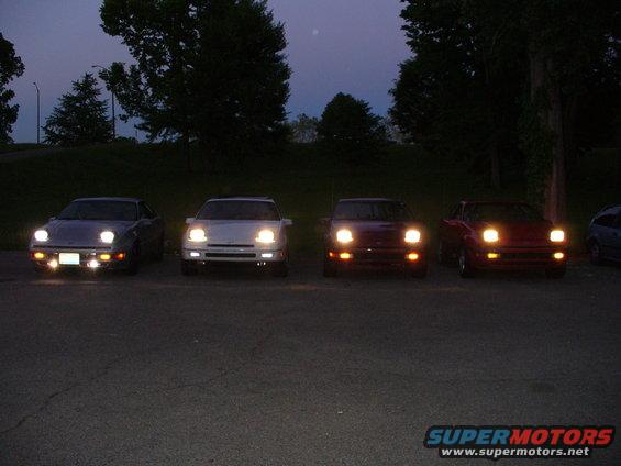 Four cars parked in a lot at dusk, all with headlights on. One is silver, two are dark red, and one is white