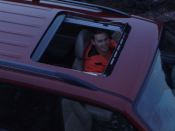 Man in orange jacket looking out of a red SUV's sunroof