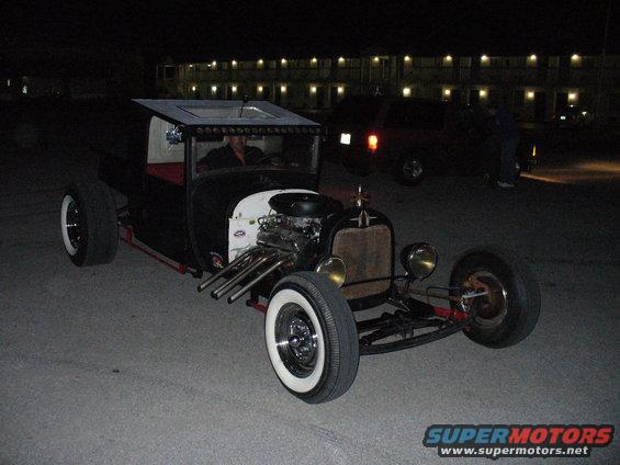 Black hot rod with exposed engine, white wall tires, and a custom roof, viewed from the front-right