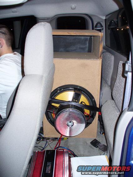 Car interior showing a large subwoofer in a wooden box, an amplifier, and a person in the driver's seat