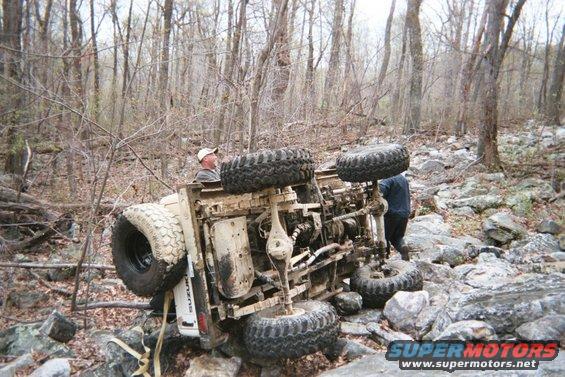Off-road vehicle flipped onto its side in a rocky, wooded area. Two people are near it