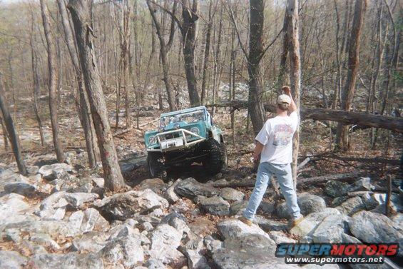1969 Ford Bronco with custom bumper and roll cage driving over rocks in a wooded area