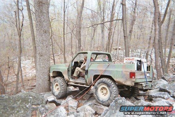 Camouflaged pickup truck with large tires on rocky terrain, driver visible inside