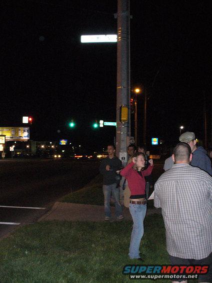 People stand on a corner at night near a street sign and traffic lights