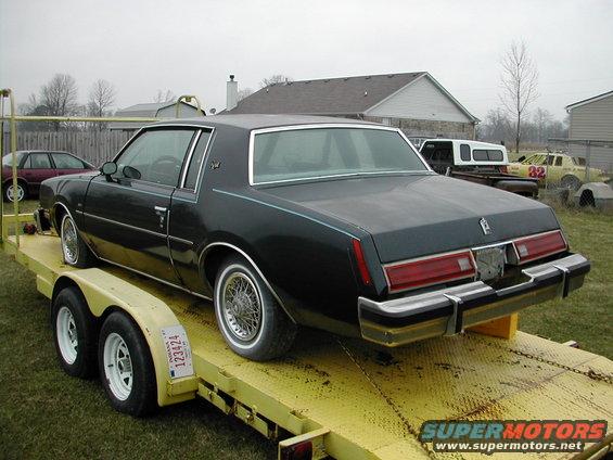 Dark sedan with wire wheels on a yellow trailer, Indiana license plate 123424 visible. Other vehicles and houses in background
