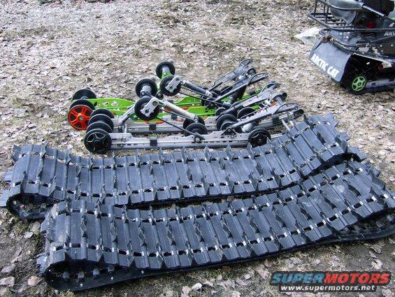 Snowmobile tracks and suspension assemblies laid out on the ground, with an Arctic Cat snowmobile in the background