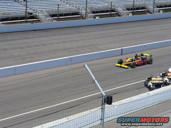 Race car with yellow and red livery on a track, with a golf cart-like vehicle nearby
