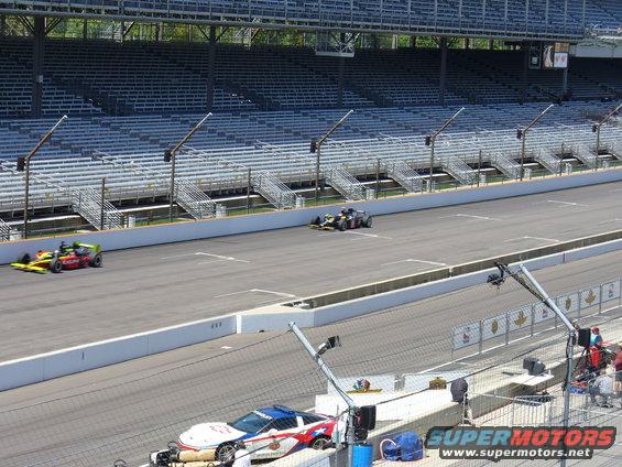 Two IndyCars race on a track in front of empty grandstands. A police car is parked in the foreground