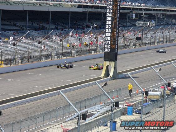 IndyCar race track with scoreboard showing lap times, multiple open-wheel race cars on track, and spectators in the stands