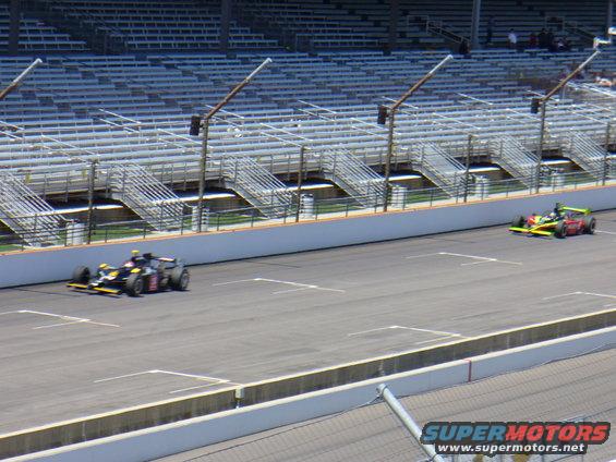 Two open-wheel race cars on a track with empty grandstands in the background