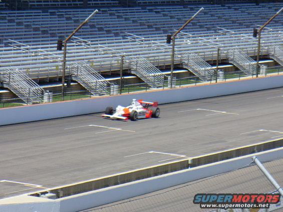 White and red open-wheel race car on a track, with grandstands in the background