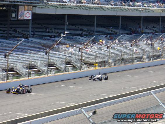 Two open-wheel race cars on a track with grandstands in the background