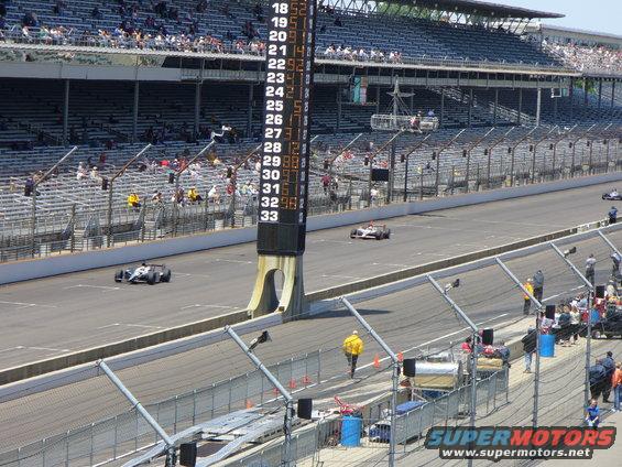 IndyCar race track with scoreboard showing lap times and multiple race cars on the track