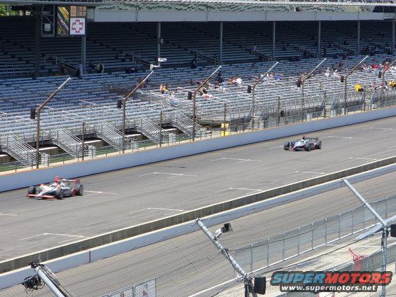 Two IndyCars race on a track with empty grandstands in the background