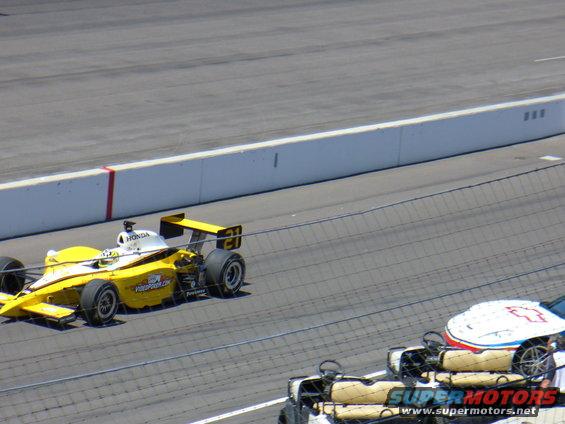 Yellow IndyCar with number 21 on track, another car partially visible in foreground