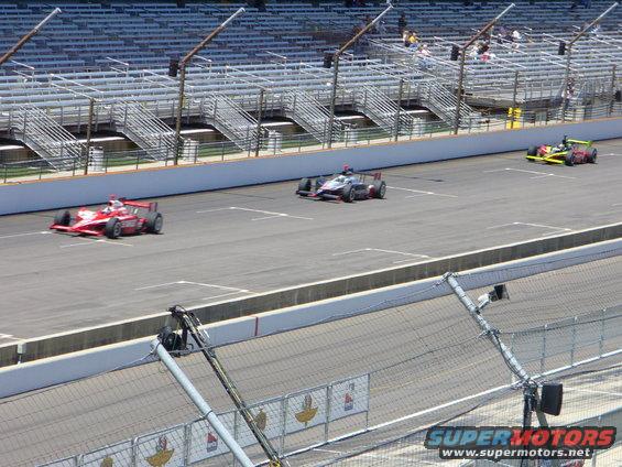 Three open-wheel race cars on a track with grandstands in the background