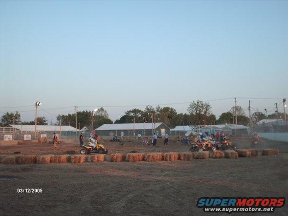 ATVs racing on a dirt track with hay bales and spectators in the background. Date stamp reads 03/12/2005
