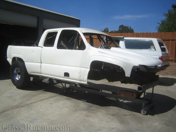 White pickup truck chassis on a stand, stripped down with a roll cage visible inside the cab