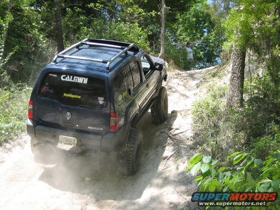 Dark blue SUV with roof rack kicking up dust on a dirt trail. 'CALMINI' sticker on rear window