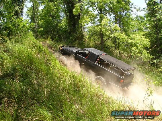 Black off-road truck with roll cage and soft top climbing a dirt hill, kicking up dust