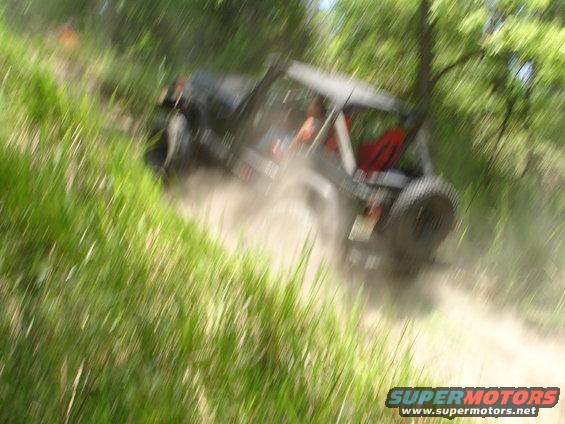 Off-road vehicle kicking up dust on a grassy hill. It has a spare tire on the back